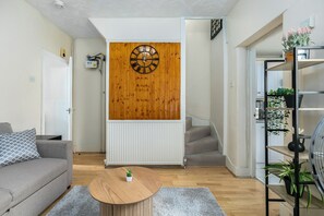 A living room with a round wooden coffee table, a grey sofa, a wooden wall clock, and a staircase leading to another room. - Rose’s Home: Charming 3-Bed House in Stratford (London)