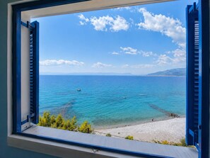 Blick auf die Küste durch ein Fenster mit blauen Fensterläden, klares blaues Meer und ein Sandstrand.
