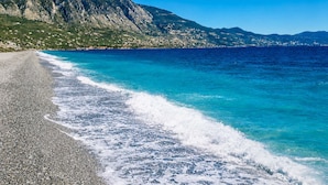 Ein Kiesstrand mit klarem, blauen Wasser und einer Bergkulisse im Hintergrund.