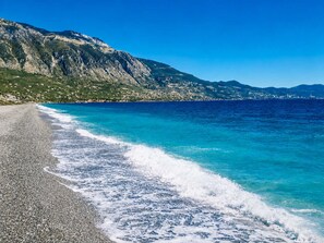 Ein Kiesstrand mit klarem, blauen Wasser und einer Bergkulisse im Hintergrund.