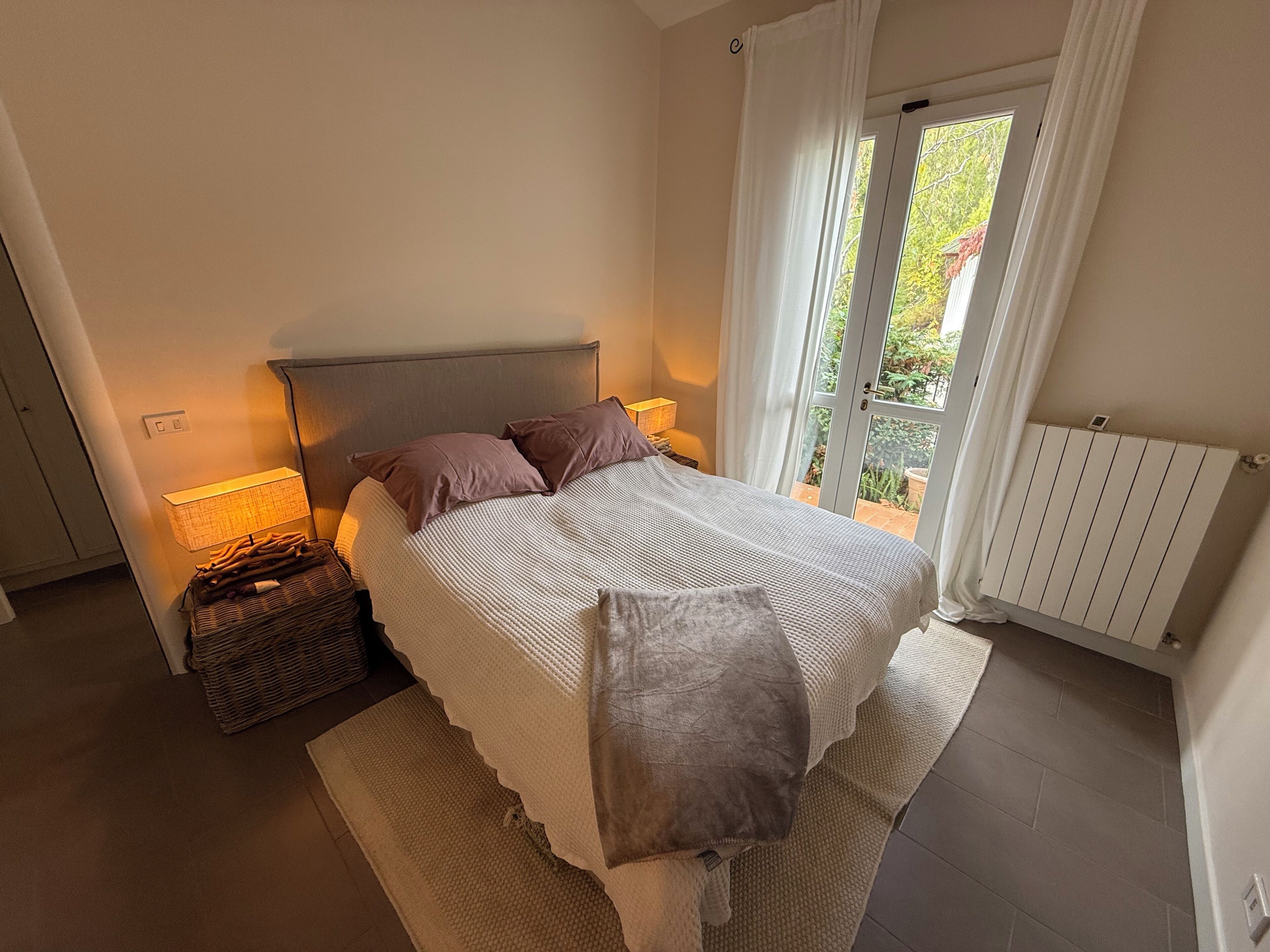A bedroom with a bed, bedside lamps, a basket, and a view through a window.