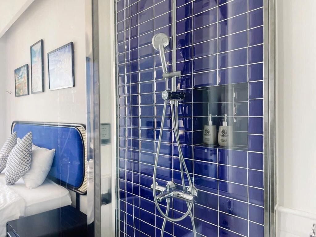 Casa de banho de hotel com zona de duche em azulejo azul, cama com cabeceira azul e um prateleira na parede com artigos de higiene pessoal.