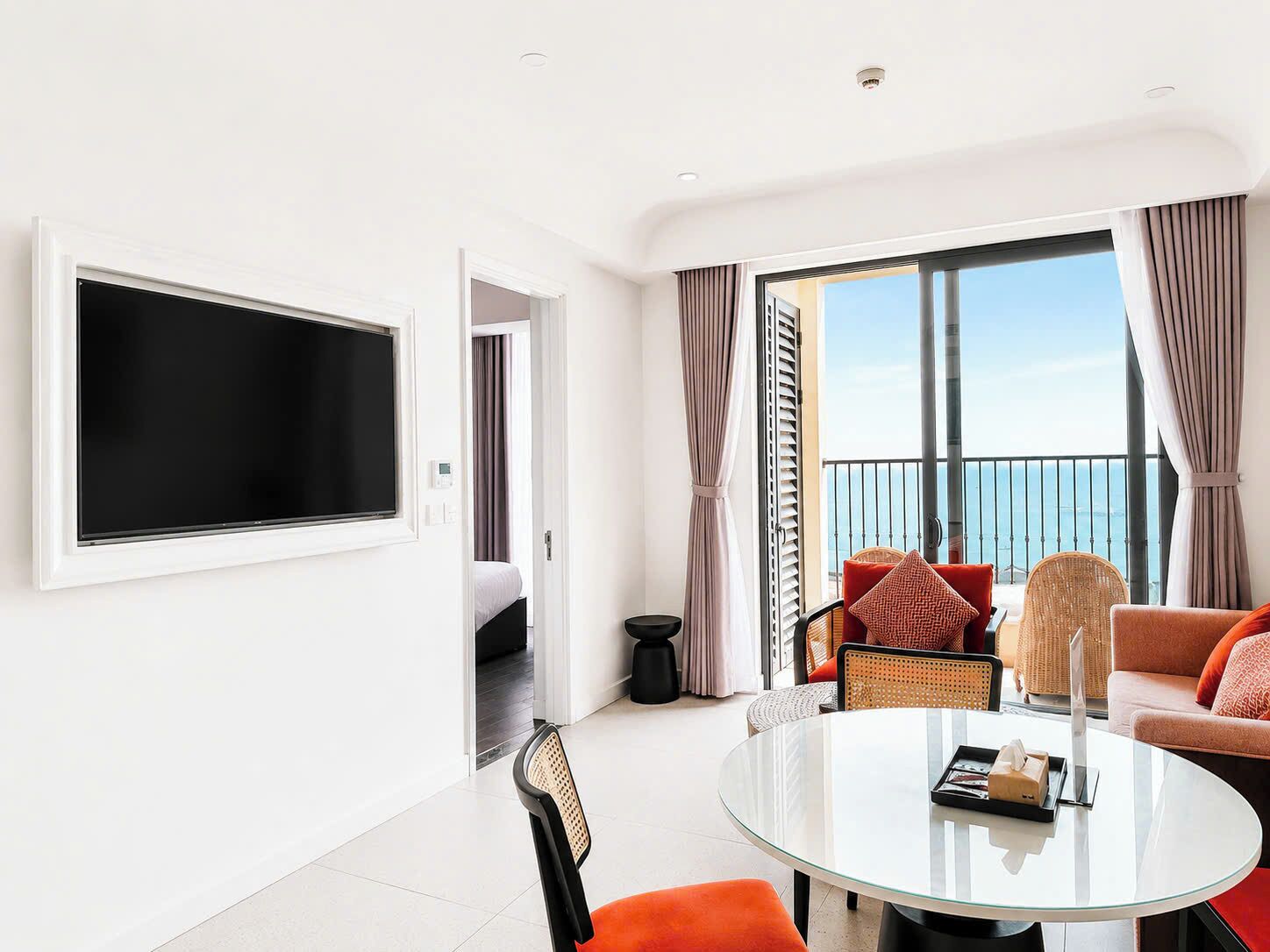 Quarto de hotel moderno com varanda, televisão de tela plana, mesa de vidro redonda e sofá com almofadas.