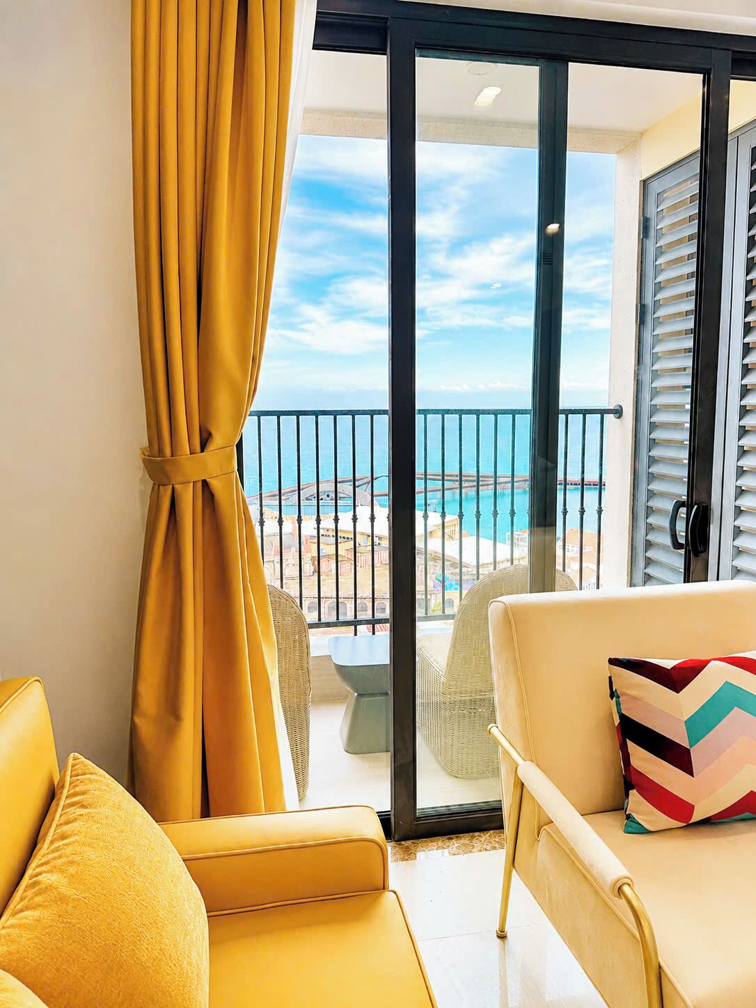 A balcony with a view of the sea and a cityscape, furnished with yellow sofas and a patterned pillow.