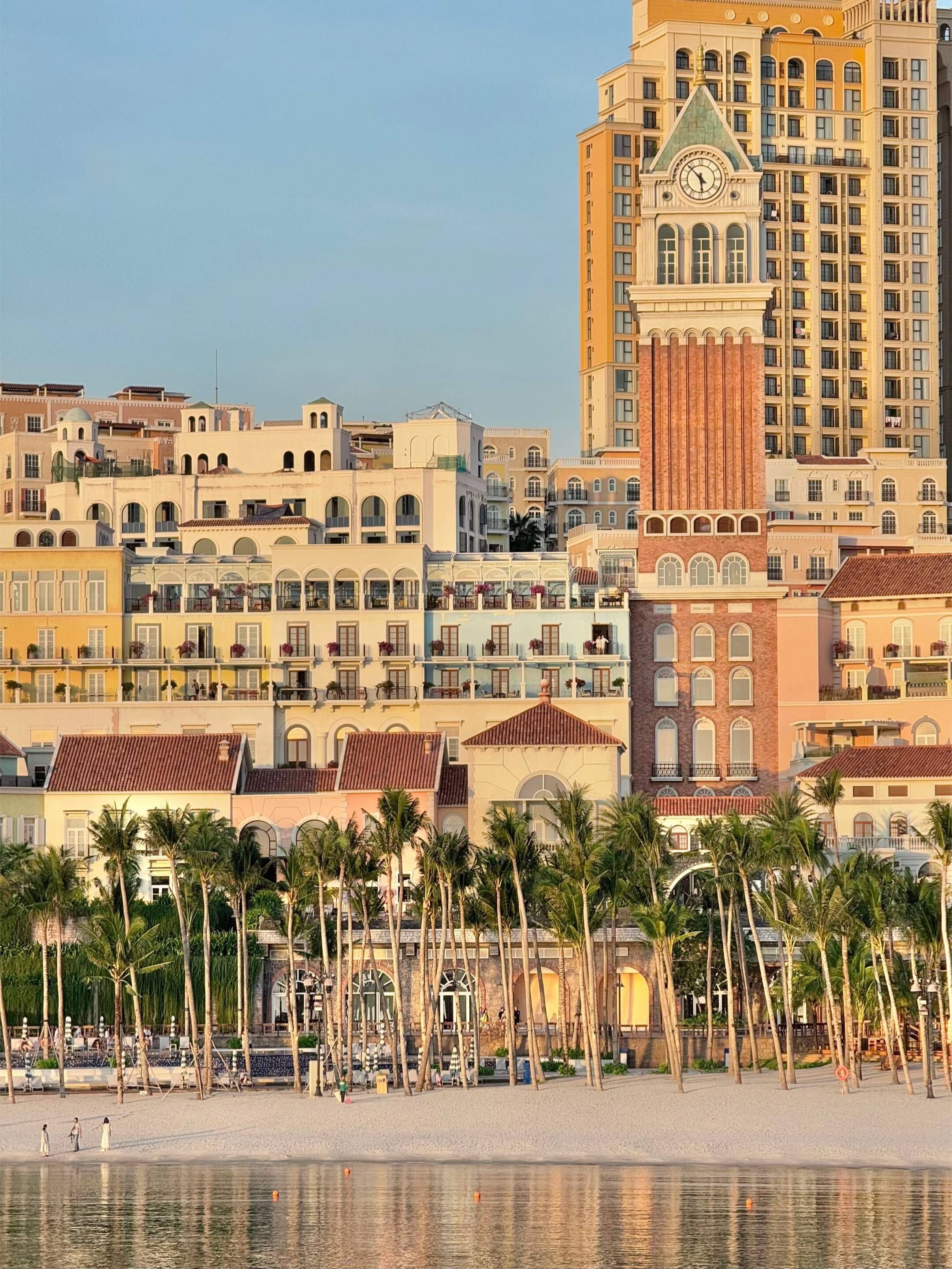 A beachfront hotel with multiple stories, a prominent clock tower, and a palm tree-lined promenade.