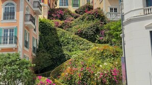 A multi-story building with balconies, a staircase with a yellow railing, and a lush garden with flowering plants.