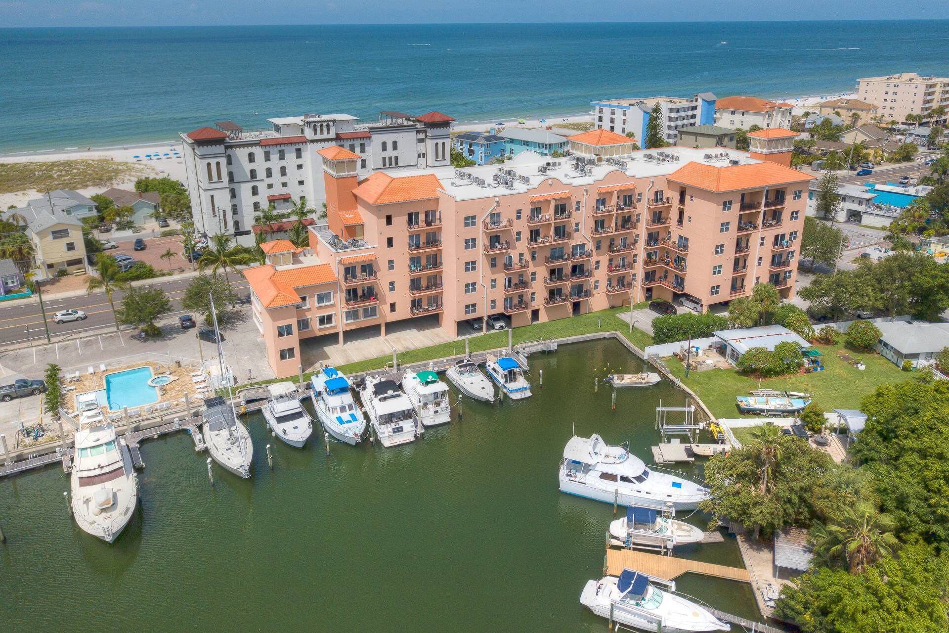 A marina with multiple boats docked, a large building with balconies, and a beach in the background.