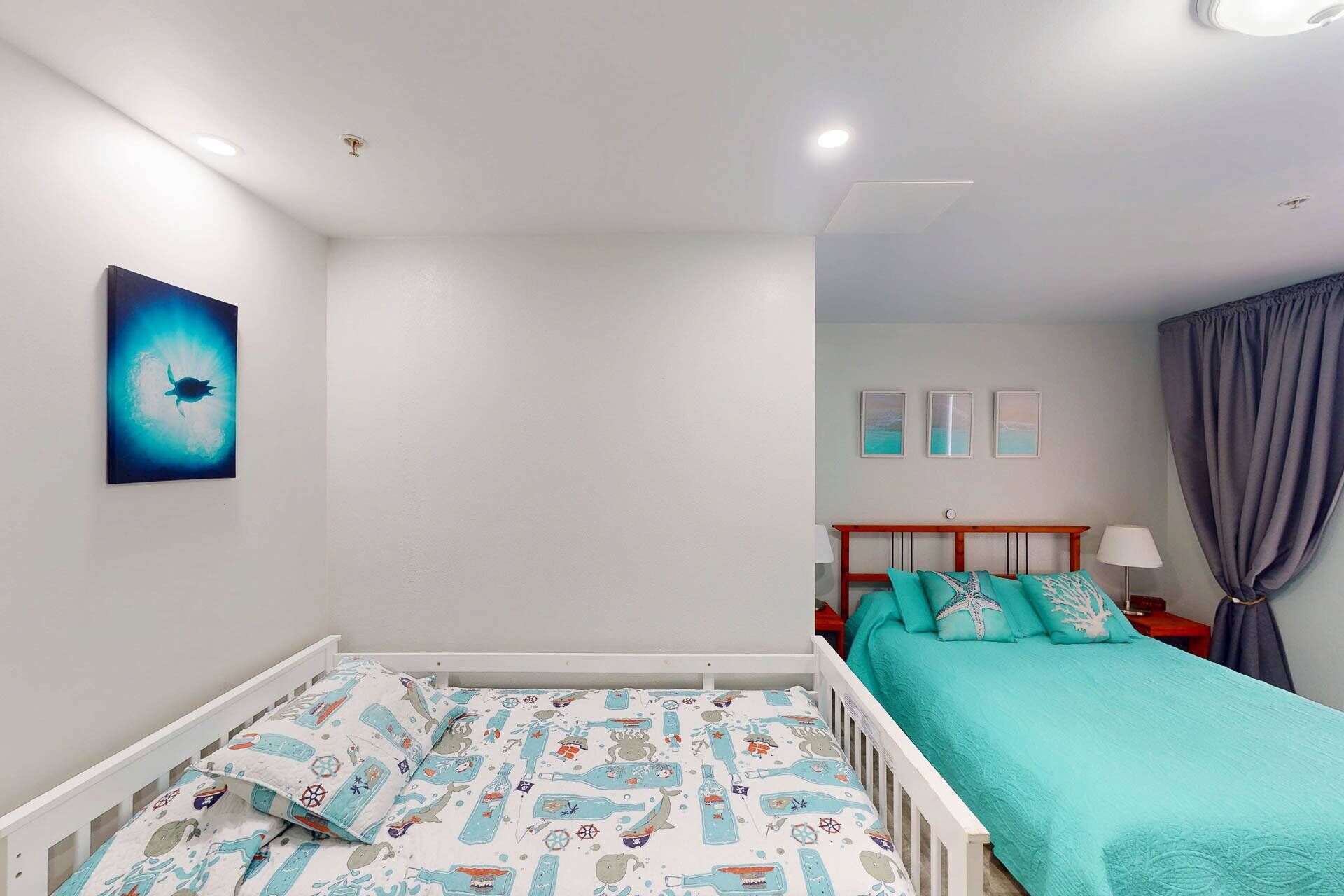 Un dormitorio con una cuna, una cama con colcha verde azulada, una mesita de noche con lámpara y una pared con tres cuadros enmarcados.