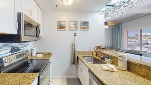 A compact kitchen with a microwave, stove, and sink. There are three framed pictures on the wall, a window with a view of boats, and a countertop with a dish soap bottle. - Private Elevator, Spectacular Pool & Marina Views, Steps to Madeira Beach & John's Pass Village (Madeira Beach)