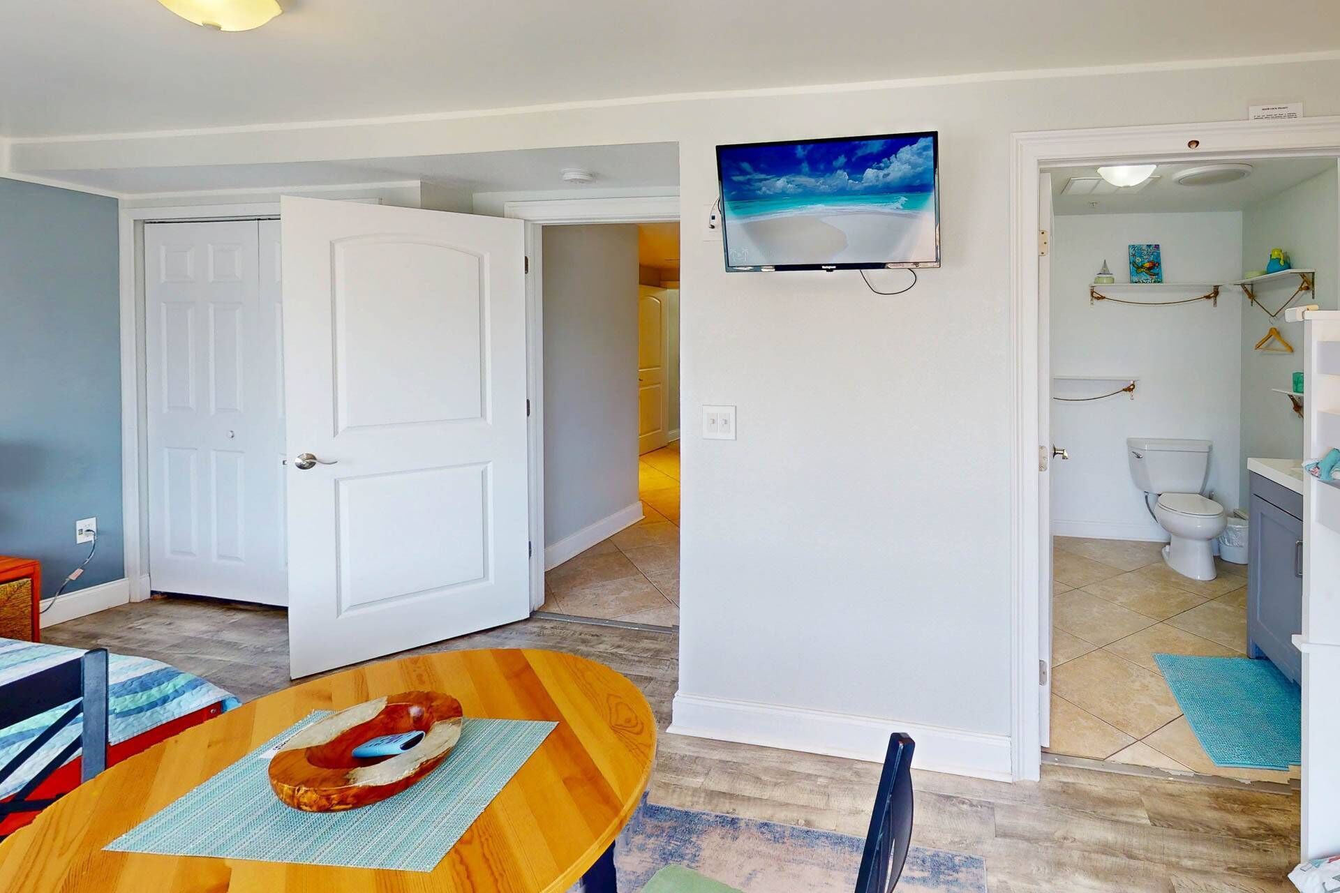 Een eetgedeelte met een houten tafel en stoelen, een witte deur en een gang naar een badkamer met toilet en planken.