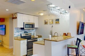 A modern kitchen with white cabinets, a stainless steel microwave, a built-in oven, granite countertops, a hanging wine rack, and a small island with a sink. - Private Elevator, Spectacular Pool & Marina Views, Steps to Madeira Beach & John's Pass Village (Madeira Beach)