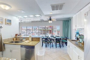 A kitchen with a dishwasher, a dining area with chairs, a ceiling fan, and a view of a marina through large windows. - Private Elevator, Spectacular Pool & Marina Views, Steps to Madeira Beach & John's Pass Village (Madeira Beach)