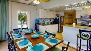 A modern kitchen with white cabinets, a central island, and a dining area with a wooden table set for six. - Private Elevator, Spectacular Pool & Marina Views, Steps to Madeira Beach & John's Pass Village (Madeira Beach)