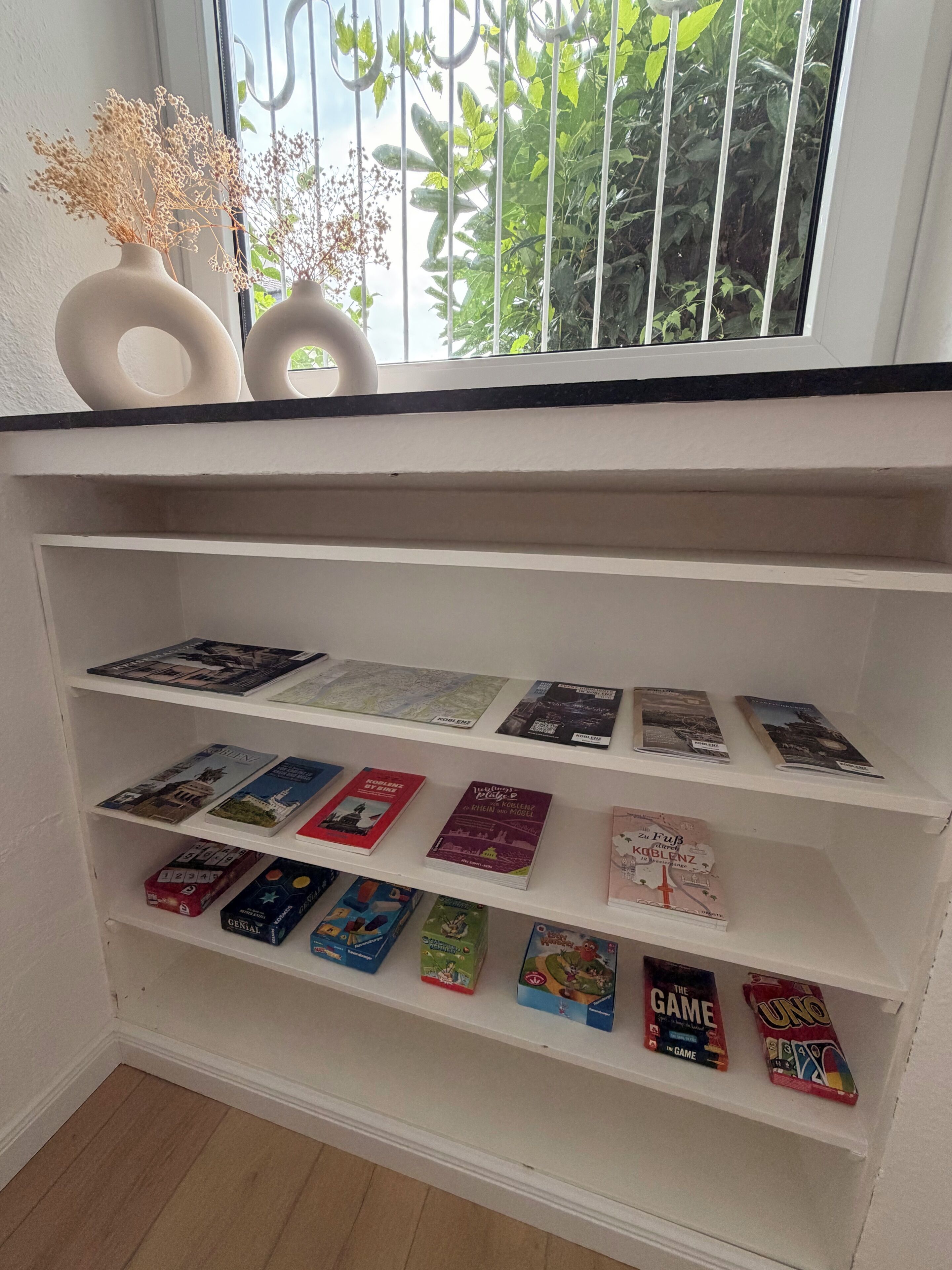 Een boekenkast met boeken en spellen, een raam met uitzicht op groen, en een decoratieve vaas met gedroogde planten.