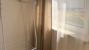 Una esquina de una habitación con un tendedero de metal, una ventana con cortinas translúcidas y vistas a un edificio.