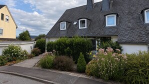 Una casa residencial con techo inclinado, un porche delantero y un jardín bien cuidado con arbustos y flores variadas.