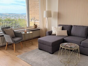 Un soggiorno moderno con un divano angolare grigio, un tavolino rotondo, una lampada da terra e vista sulla città attraverso ampie finestre.