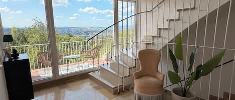 A spacious room with a view of a cityscape through large windows, a staircase, and a seating area with chairs and a table.