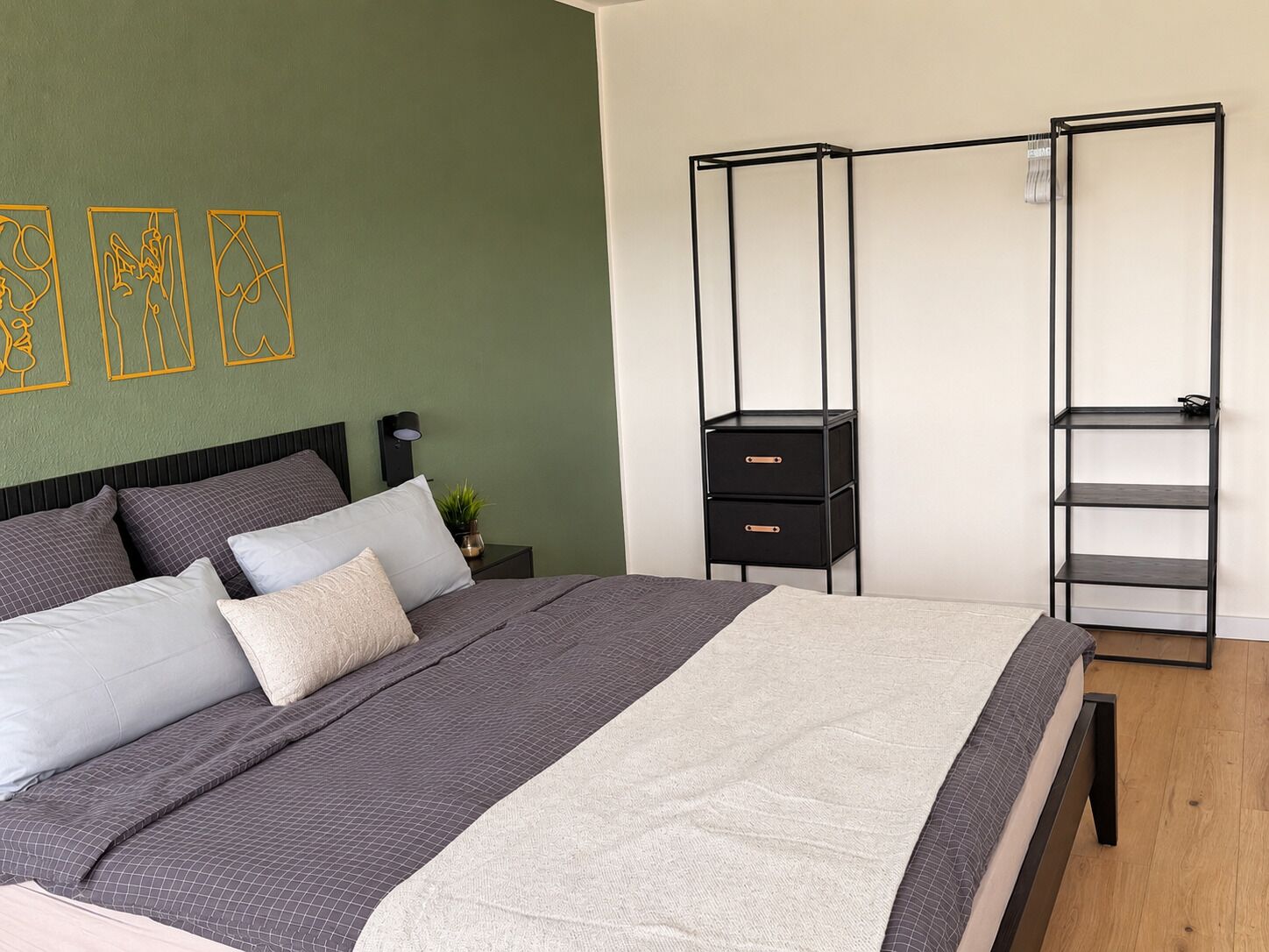 Una camera da letto con un letto, un comodino, un contenitore in metallo nero con cassetti e una mensola a parete.