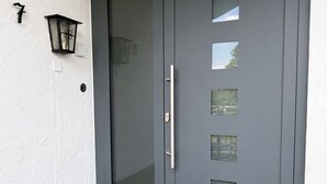 Puerta doble de color gris con manija y paneles de vidrio esmerilado, felpudo negro y lámpara de pared.