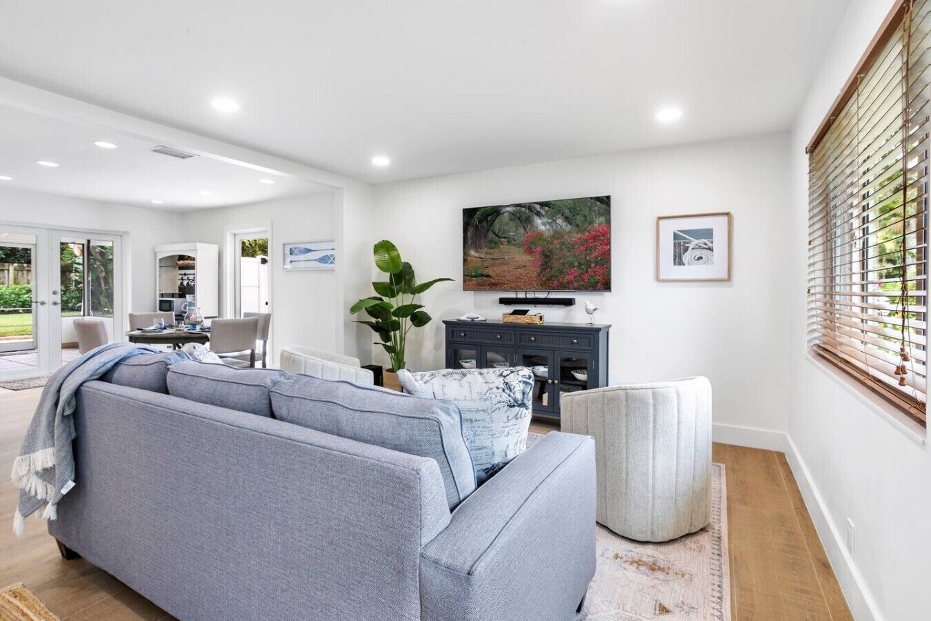 Ein modernes Wohnzimmer mit grauem Sofa, einem Couchtisch und einem Fernseher. Ein großes Fenster mit Jalousien, eine Pflanze und Kunstwerk sind an der Wand zu sehen.