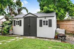 A small, single-story shed with a dark door and windows, surrounded by a grassy yard and a wooden fence. - 3BD/2BA Palm Gardens Retreat (Palm Beach Gardens)