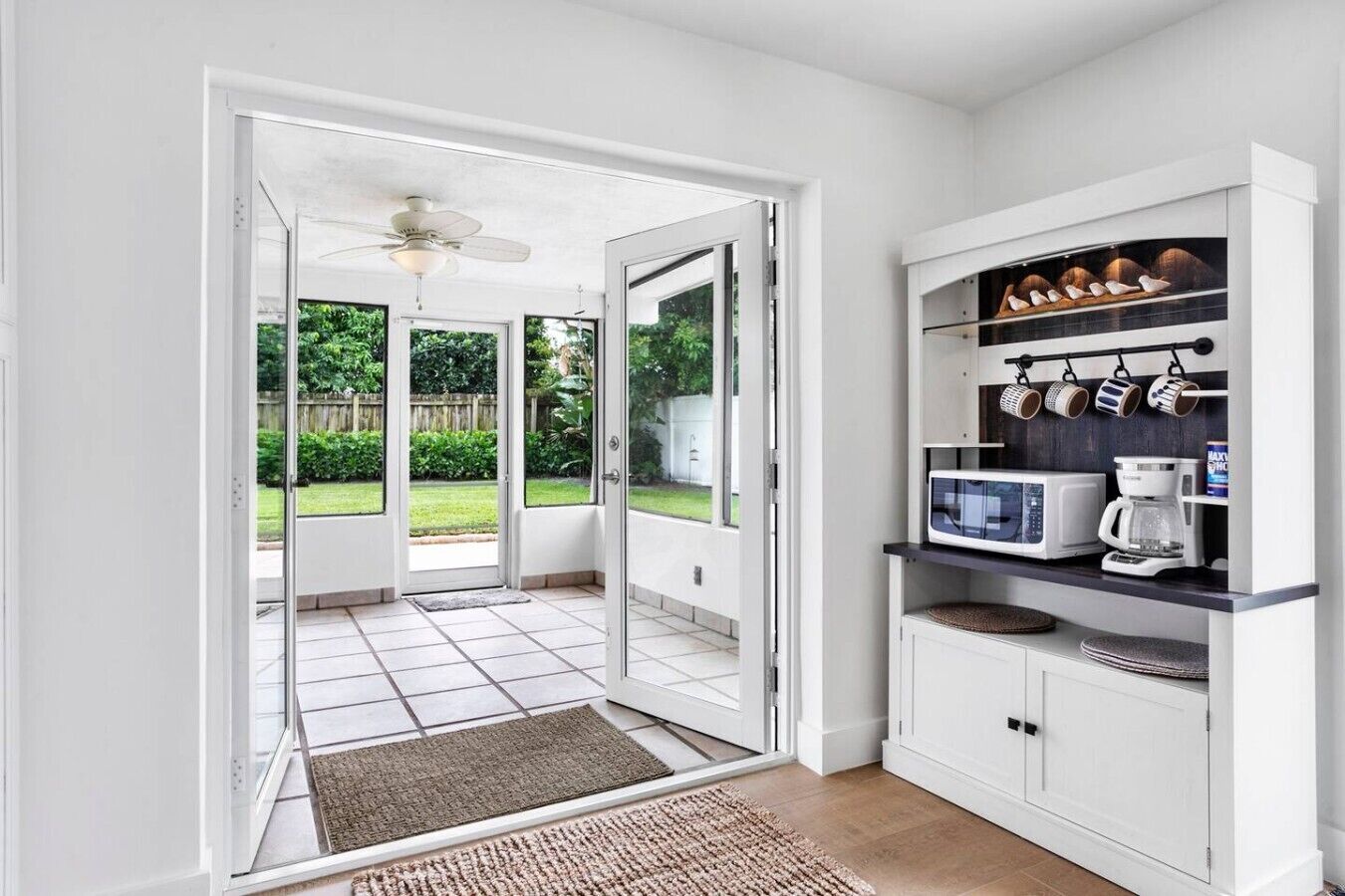 An open door leading to a patio with a ceiling fan, a kitchen area with a microwave, coffee maker, and shelves with mugs.