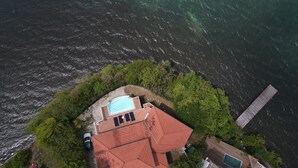 Ett flygbildsfoto av en fastighet vid kusten med en swimmingpool, en byggnad med rött tak och en träbrygga som sträcker sig ut i vattnet.