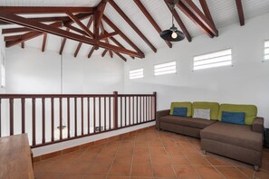 Ein geräumiges Zimmer mit hoher Decke, einem Holzbalkon und einem braunen Sofa mit Kissen sowie einem Fliesenboden.