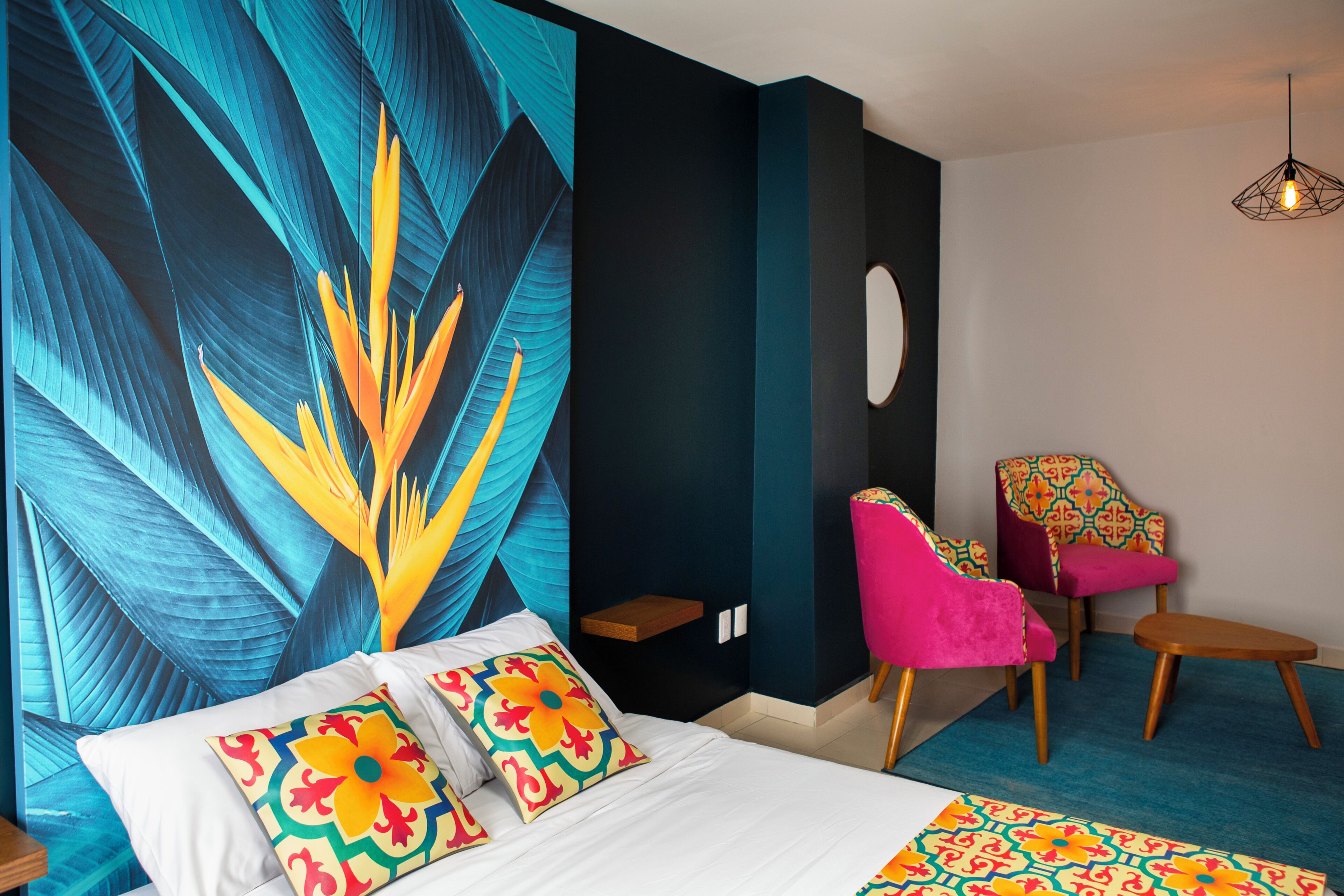Habitación de hotel con un gran mural en la pared, una cama con almohadones de colores, una silla con cojín estampado y una lámpara de techo original.