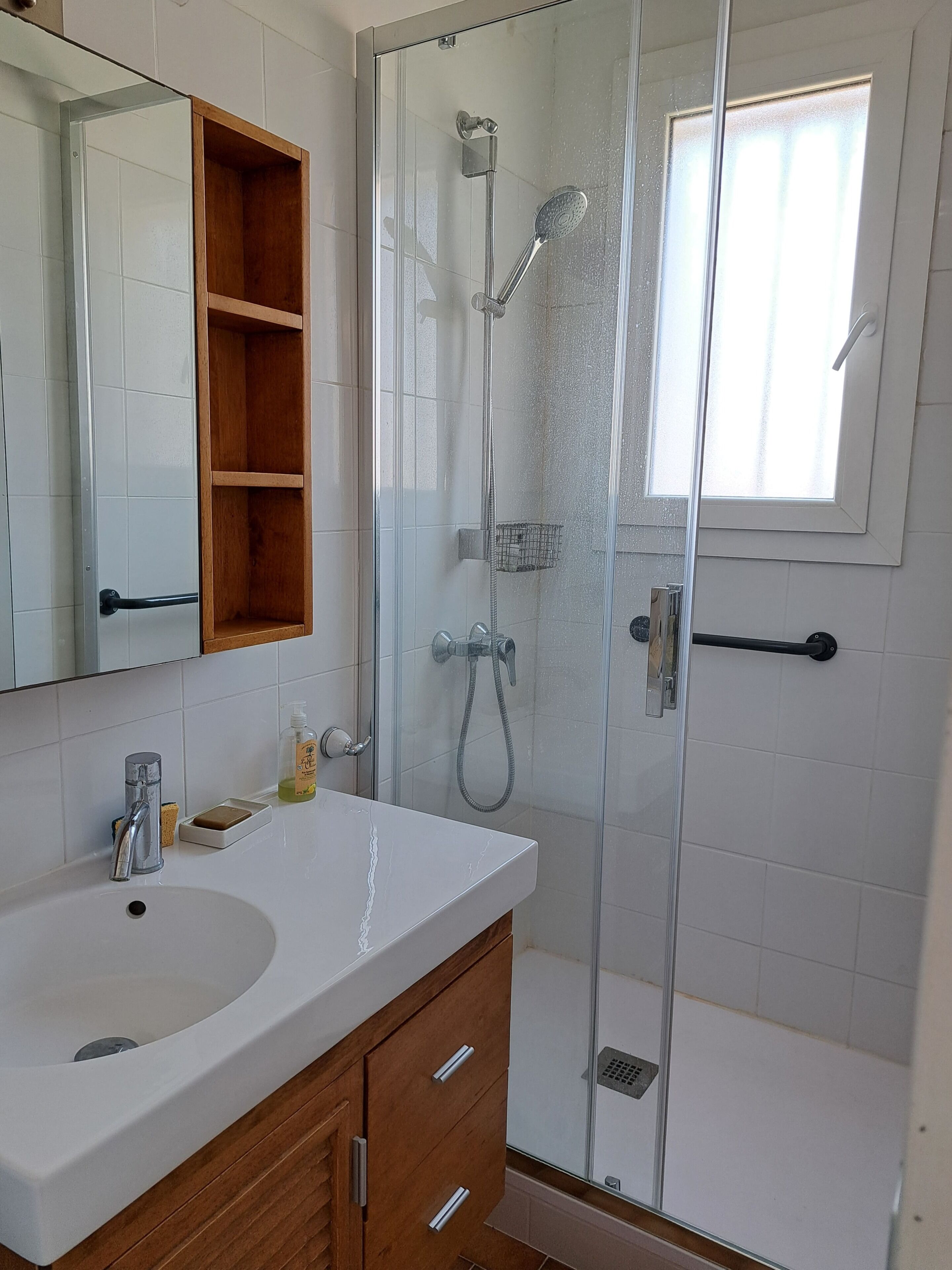A bathroom with a white sink, wooden vanity, a glass shower enclosure, and a window.