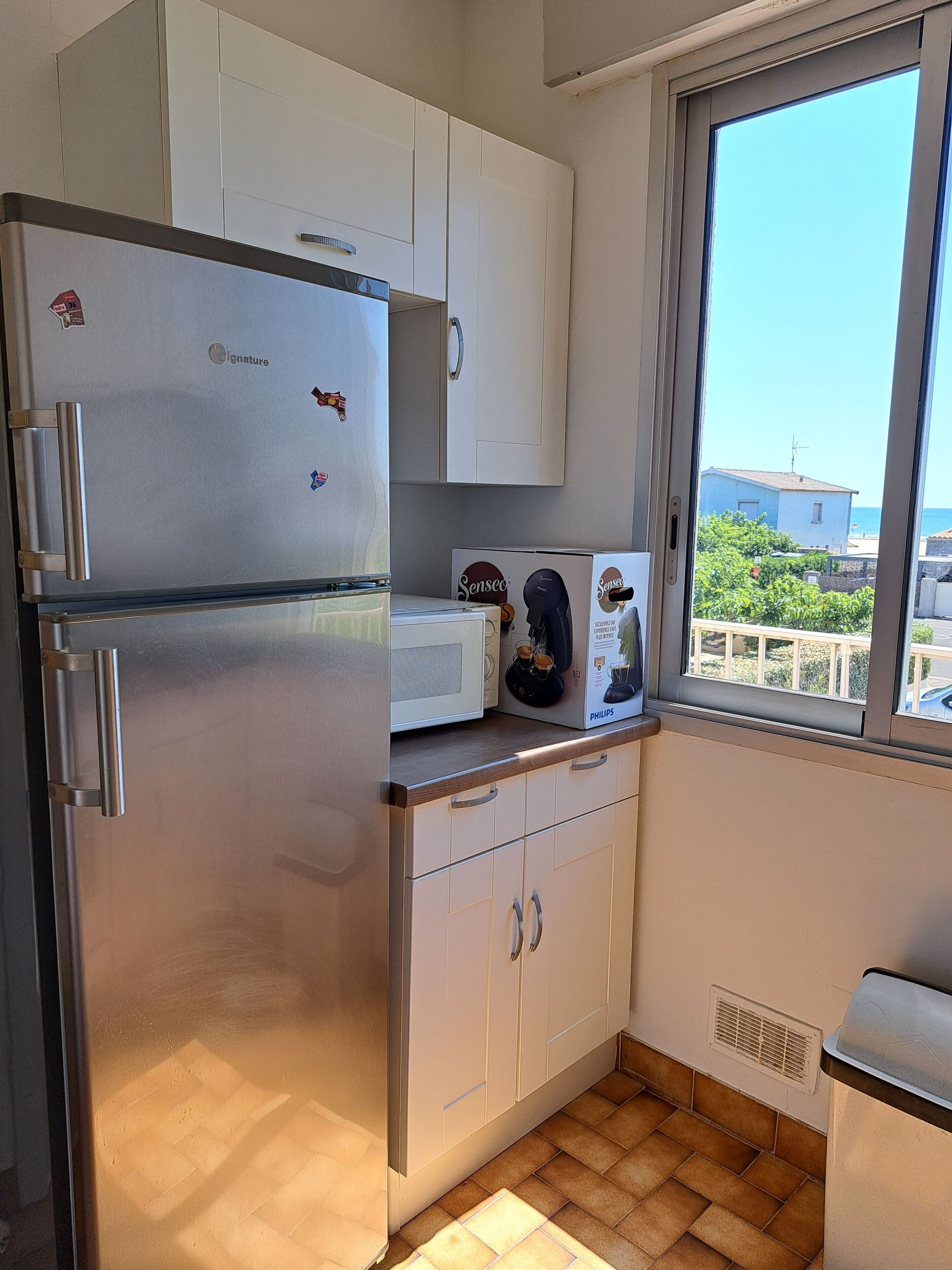 Cocina compacta con refrigerador de acero inoxidable, microondas y ventana con vistas a edificios y zonas verdes.