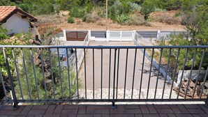 A balcony with a metal railing, a paved area, and a view of a road with a bench and a car.