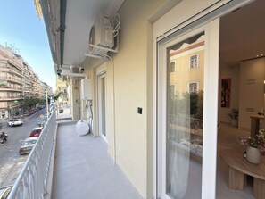 A balcony with a view of a busy street, a window with a view of a building, and a small table with a vase. - Stylish City 2BR Apartment | Ad Residences (Thessaloniki)
