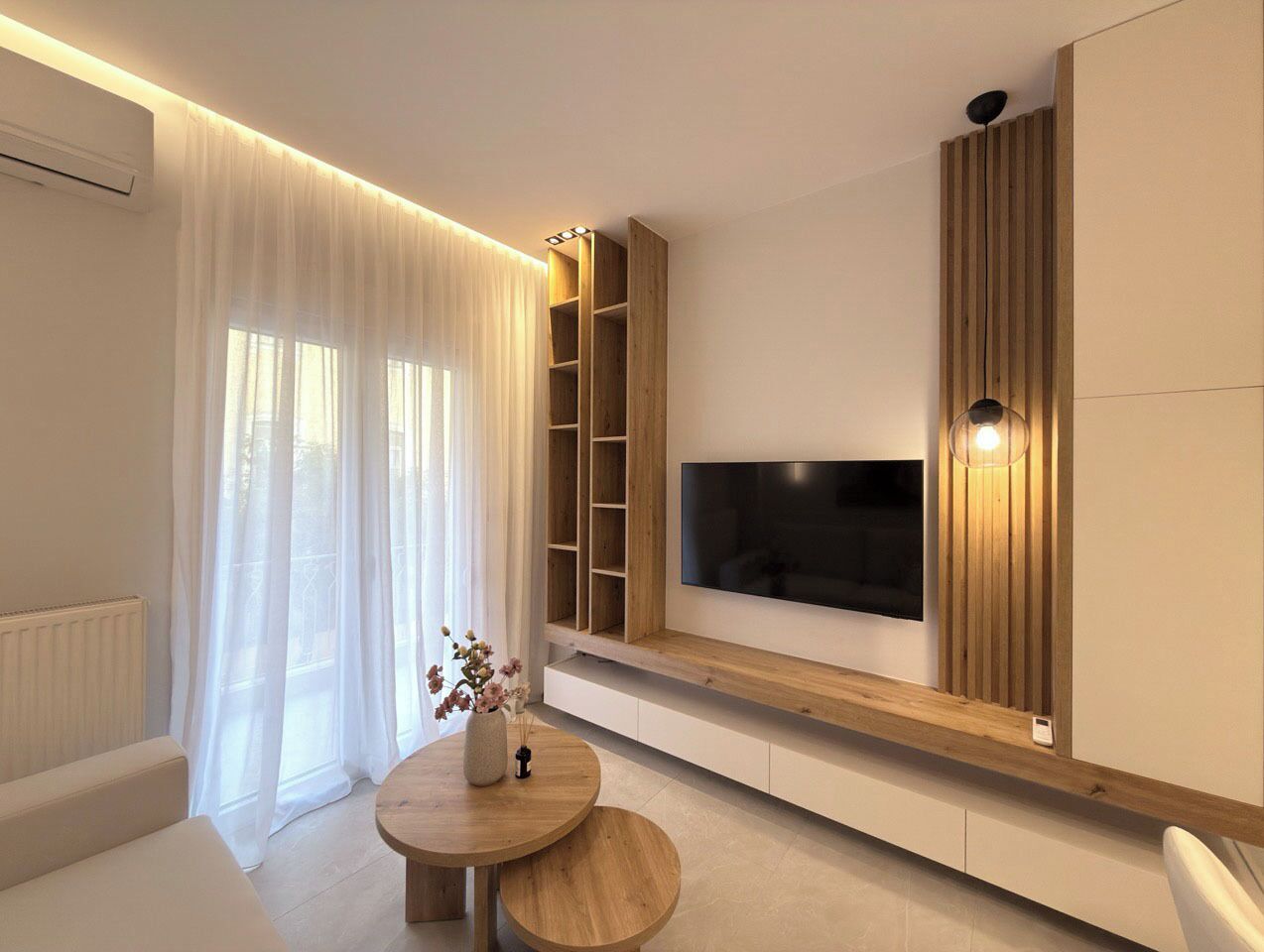 Un salon moderne avec un téléviseur à écran plat fixé sur une étagère en bois, une table basse ronde et un canapé blanc.