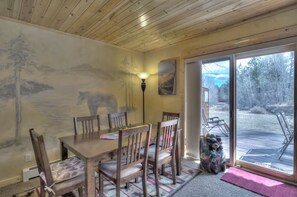 A dining area with a table and chairs, a wall mural, a lamp, and a view of a deck and outdoor area. - Bear Creek TWO by Notable Vacation Rentals (Estes Park)