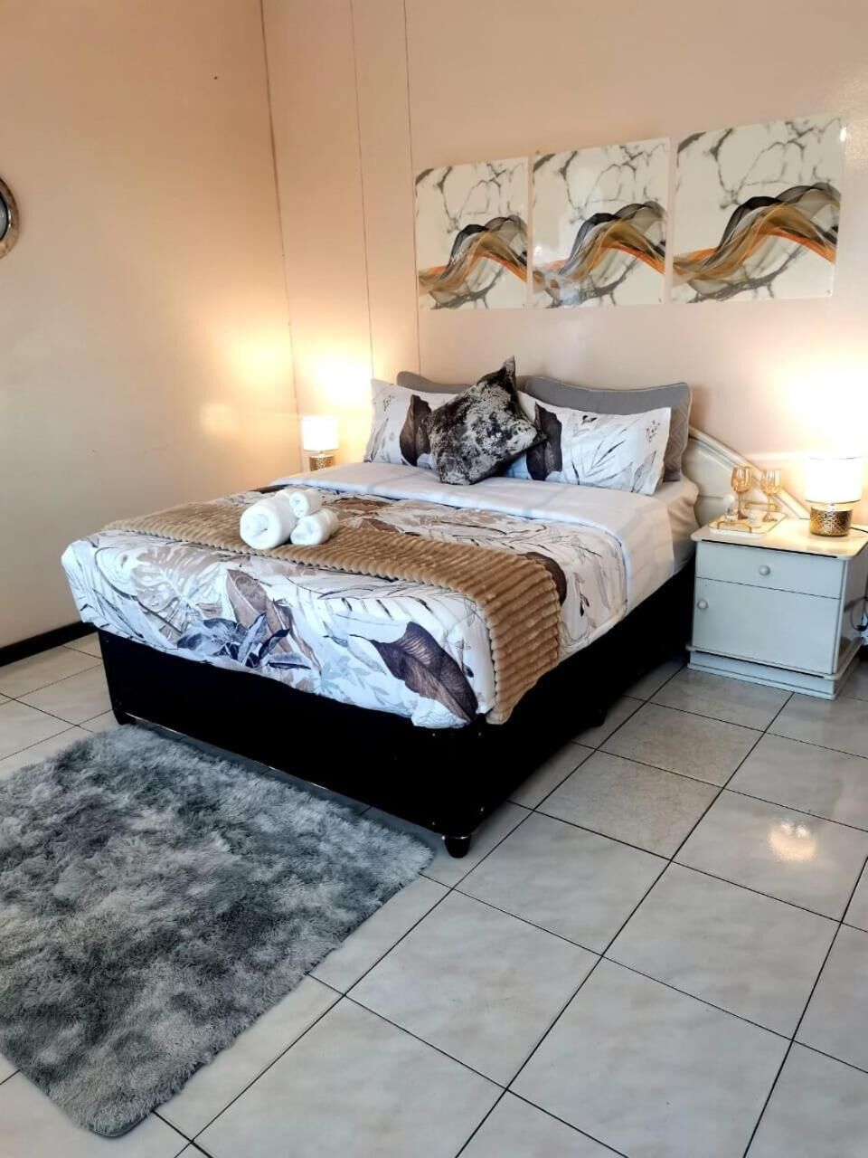 A neatly made bed with a patterned bedspread, a grey fluffy rug, and a bedside table with a lamp.