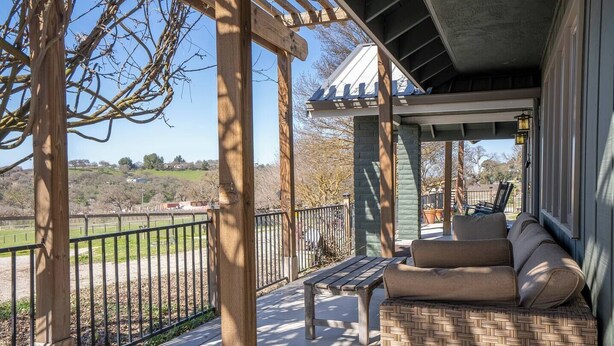 A covered outdoor seating area with a wooden bench and a woven table, overlooking a fenced property. - Harvest Ridge Ranch 5 Bedroom Farmhouse Sleeps 14 (Paso Robles)