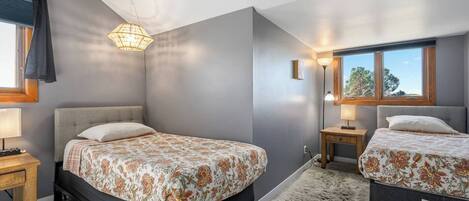 A bedroom with two beds, a chandelier, a window with a view, and a wooden nightstand.