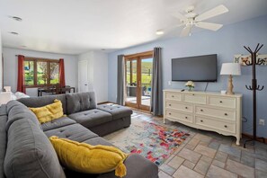 Un salon avec un canapé d’angle gris, une commode blanche, un téléviseur à écran plat et un ventilateur de plafond.