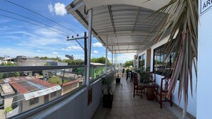 Un balcón techado con vistas a edificios, plantas en macetas y mobiliario de exterior.