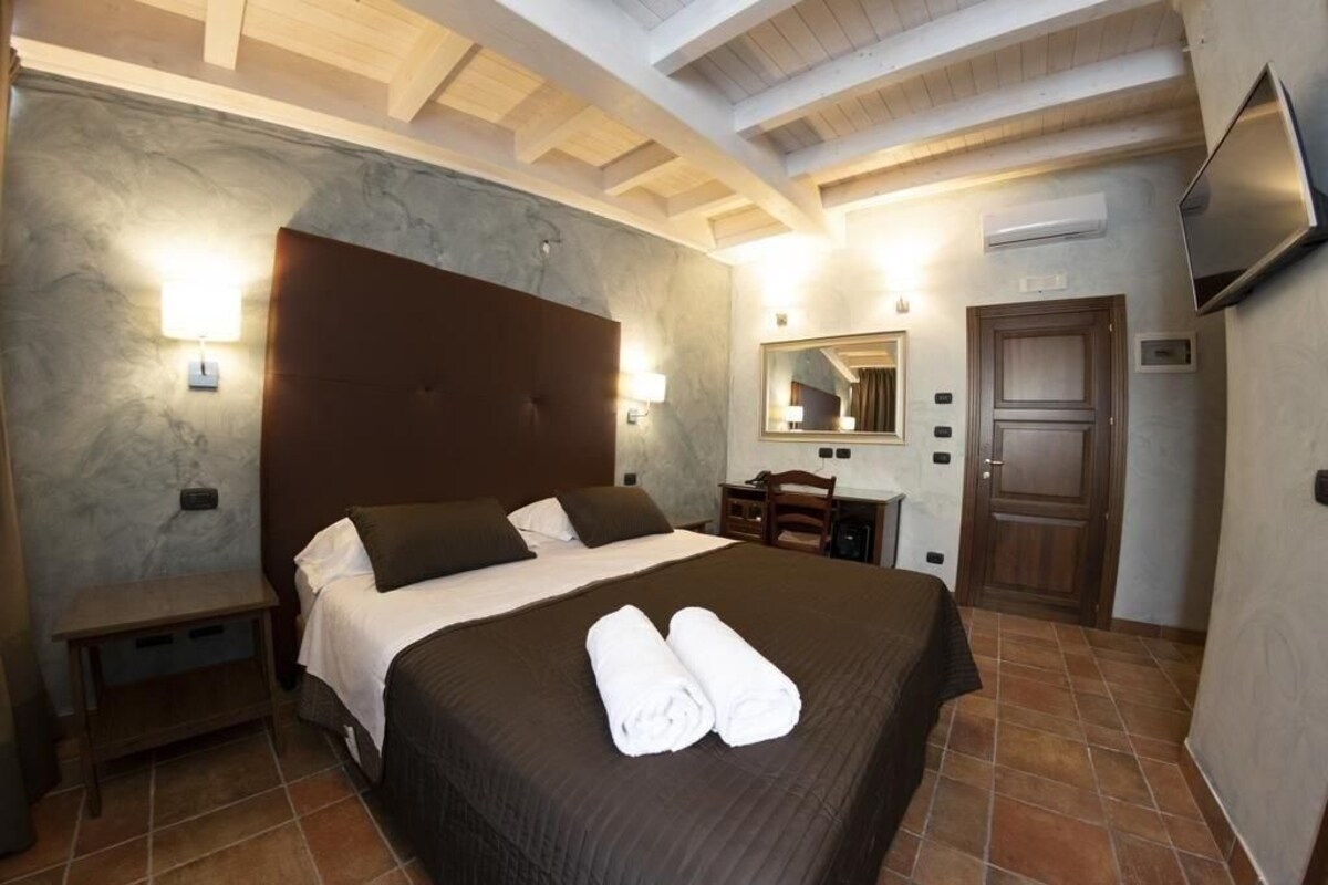 A hotel room with a large bed, a wooden headboard, a bedside table, a desk, a chair, a mirror, and a TV.