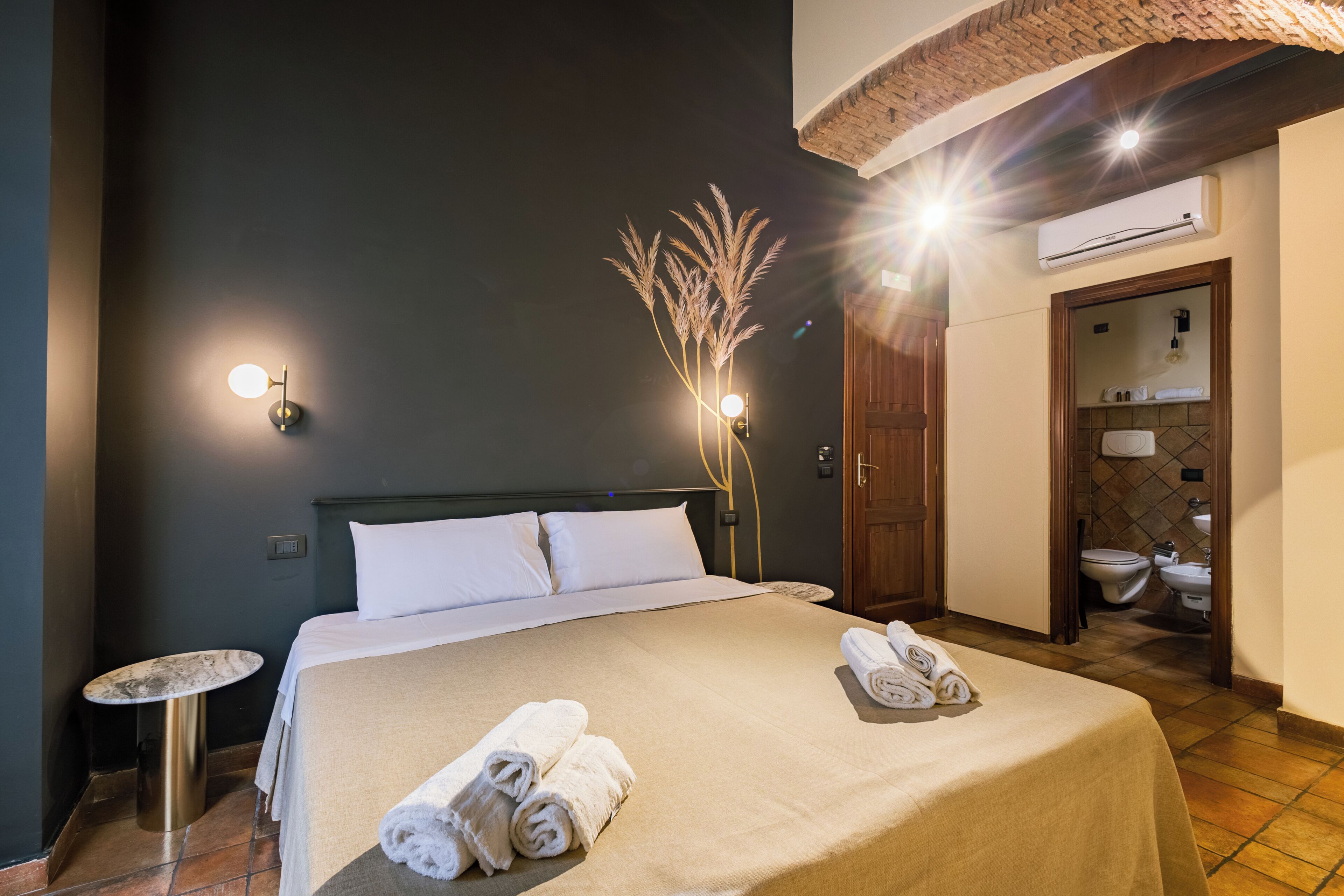 A hotel room with a bed, a bedside table, a wall-mounted lamp, and a bathroom visible through an open door.
