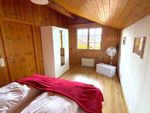 Ein Zimmer mit Holzwänden und schräg versetzter Decke, ein Bett mit roter Decke, ein weißer Kleiderschrank, ein Heizkörper und ein Fenster mit Aussicht.