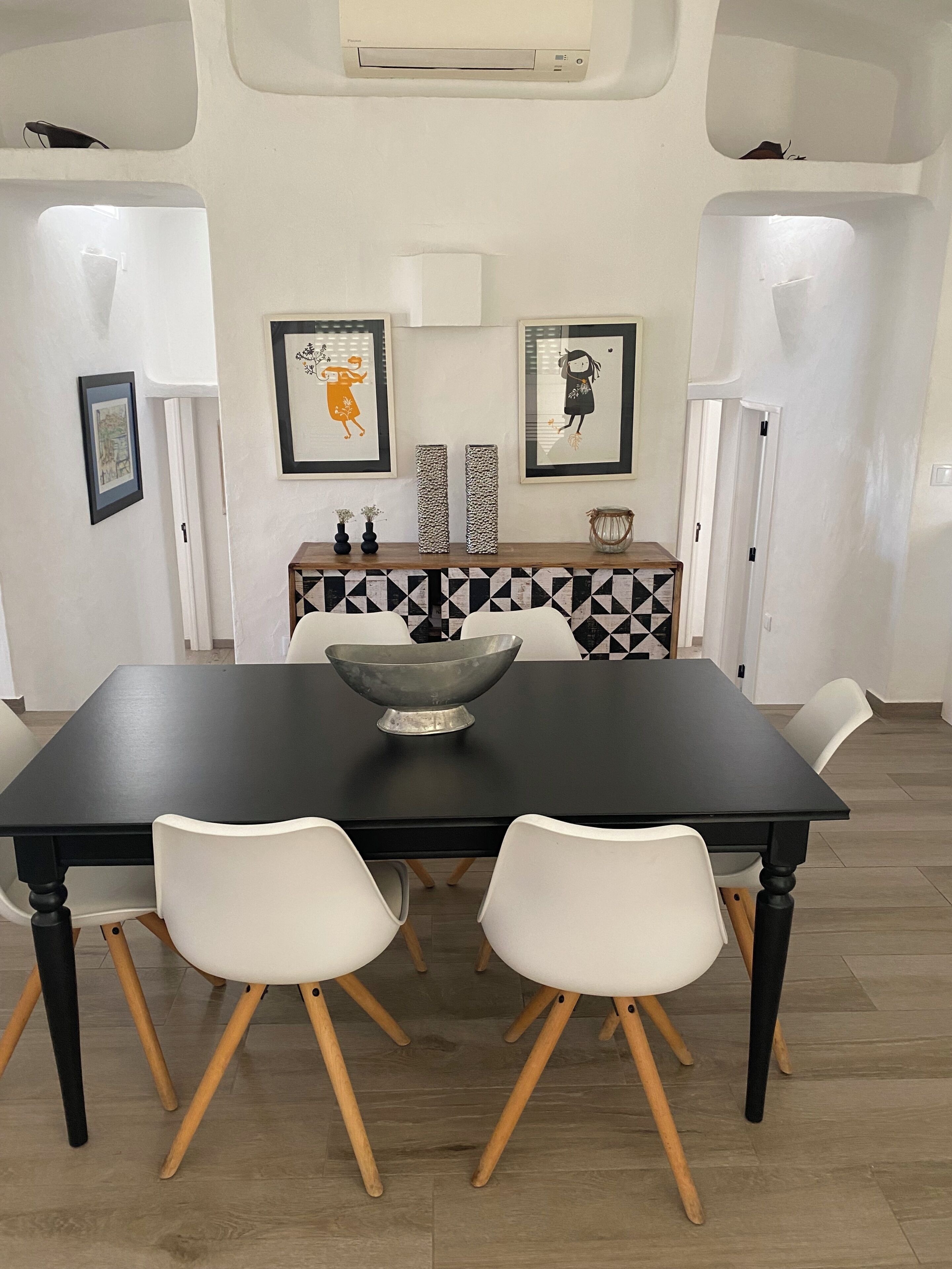 Un comedor con una mesa negra, sillas blancas, un cuenco decorativo y dos jarrones sobre un mueble estampado.