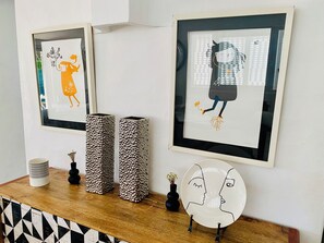 Zwei gerahmte Kunstwerke an einer Wand, eine Vase und ein Teller mit einem Gesichtsmotiv auf einer Holzoberfläche.