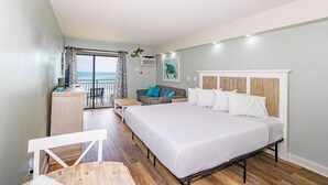 A hotel room with a large bed, a desk, a chair, a balcony with a view, and a painting on the wall. - Sea Mist 20509 Direct Ocean Front Condo (Myrtle Beach)