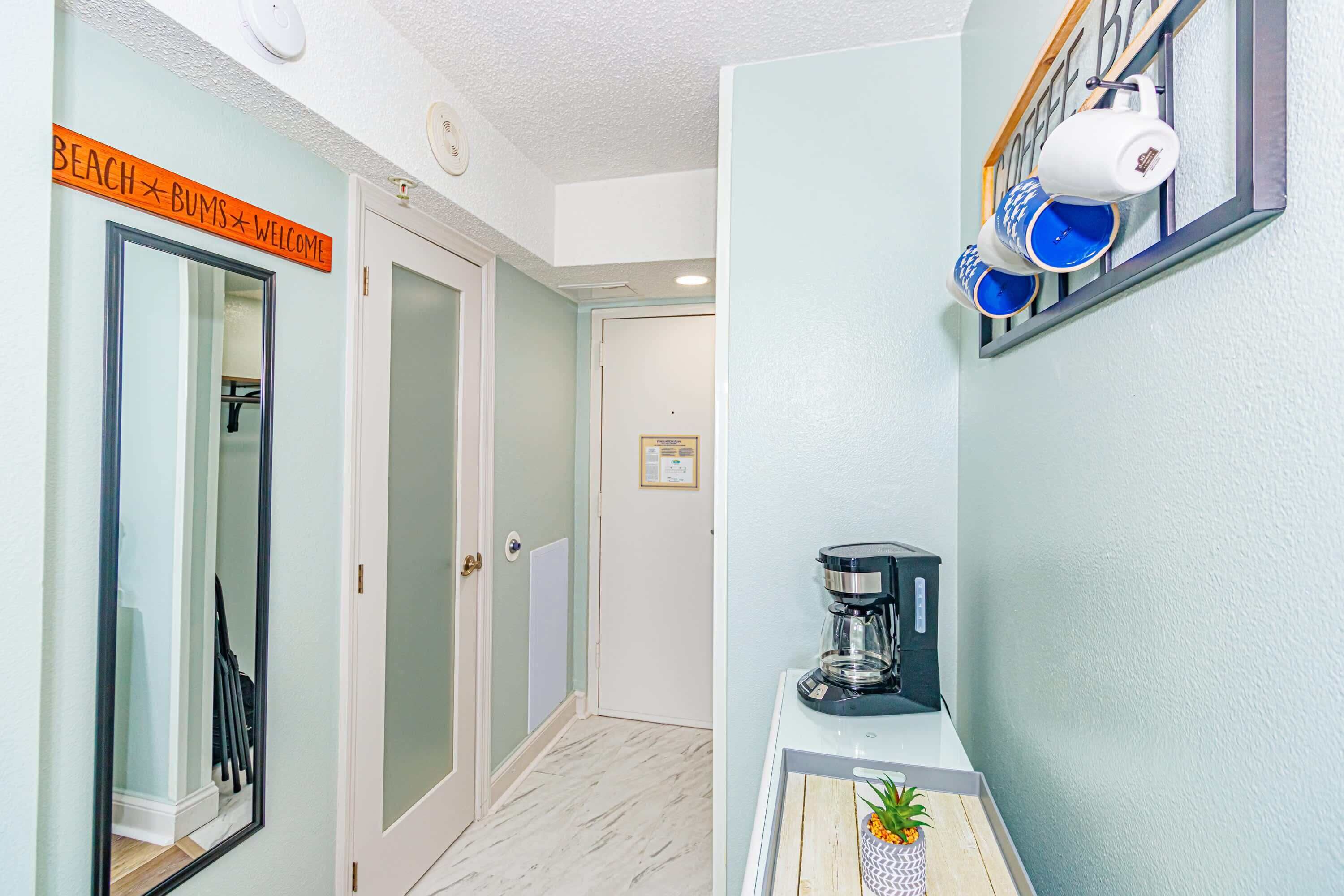 A hallway with a coffee maker on a table, a sign reading 'BEACH BUMS Welcome', and a coffee machine on the wall.