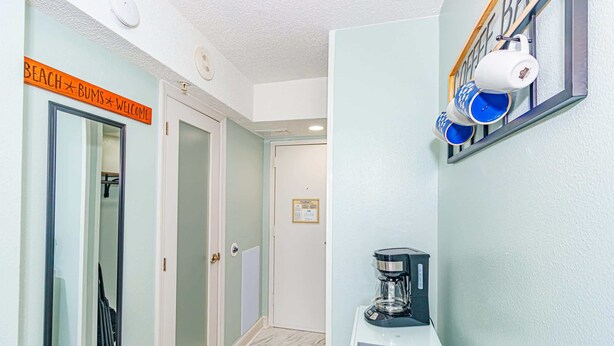 A hallway with a coffee maker on a table, a sign reading 'BEACH BUMS Welcome', and a coffee machine on the wall. - Sea Mist 20509 Direct Ocean Front Condo (Myrtle Beach)