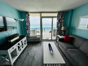 A living room with a sofa, a coffee table, a TV, and a view of the ocean through large windows. - Paradise by the Sea. Oceanfront. (Myrtle Beach)